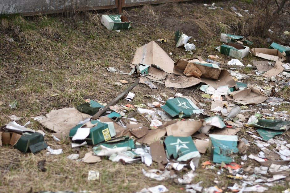 An outdoor area with scattered rubbish including broken cardboard boxes, crushed paper, and discarded packaging materials across a grassy patch near a concrete barrier. Several flattened green cardboard boxes with white star logos are visible, some torn or partially smashed. Small pieces of paper and plastic waste are mixed within the debris, which also includes a bent metal rod lying among the trash. The environment appears untidy and unmanaged, with litter spread unevenly over the ground, indicating an instance of illegal dumping or neglected waste collection. The background shows a slight incline with dry, sparse grass and some leafless shrubbery, suggesting an urban or suburban setting. This scene exemplifies the importance of proper rubbish removal services, such as those offered by Rubbish Clearance Harrow, to manage waste properly and maintain environmental cleanliness.