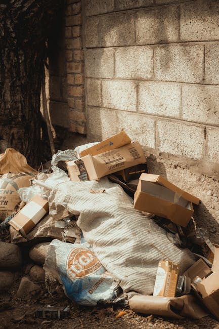 A close-up of a person wearing a light pink glove, holding a transparent plastic bag filled with crushed and empty plastic bottles of various sizes and shapes. The bottles are clear with visible ridges and caps still attached. The background features a plain, light yellow curtain or wall, providing a neutral setting. The scene emphasizes the collection and disposal of plastic waste, relevant to waste management and rubbish removal services. The lighting is even, highlighting the glossy surfaces of the plastic bottles and the clarity of the bag, with no shadow distractions. This image illustrates the process of gathering recyclable plastic materials, supporting the context of private disposal and environmentally conscious rubbish clearance efforts undertaken by companies such as Rubbish Clearance Harrow, especially in relation to alternative waste handling methods outside of local authority collection.