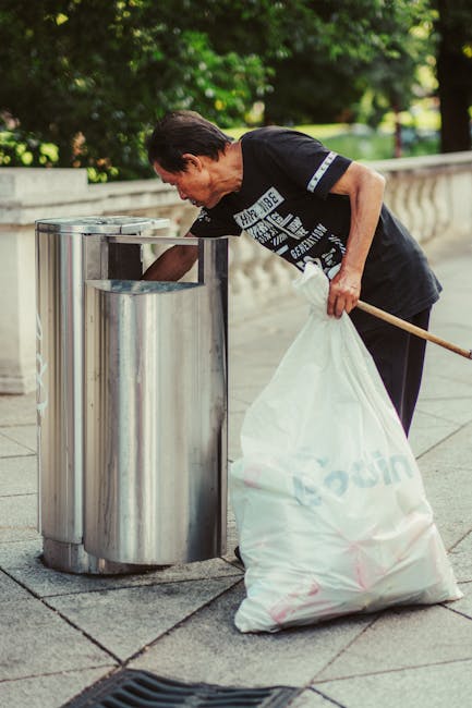 A middle-aged man with dark hair, wearing a black T-shirt with white text and dark trousers, is seen leaning forward as he disposes of rubbish into a cylindrical stainless steel public waste bin located on a paved sidewalk. He holds a long-handled tool in one hand and a large white plastic bag filled with collected waste in the other, which appears to contain various types of rubbish. The surrounding environment includes greenery with trees in the background, and a stone balustrade along the edge of the sidewalk suggests a public park or promenade area. Natural daylight illuminates the scene, highlighting the reflective surface of the waste bin and the textures of the paving stones. The image captures an act of private waste disposal, typical of independent rubbish clearance, aligned with rubbish removal services offered by Rubbish Clearance Harrow and similar providers.