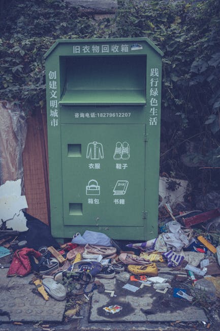 A green, weathered clothing donation bin located outdoors on a pavement beside dense shrubbery and tree foliage. The bin has a large rectangular opening at the top for depositing items, with Chinese characters above indicating it is for old clothing recycling. Below the main opening, there are smaller compartments labeled in Chinese for specific item types, including clothing, shoes, bags, and books, with symbols representing each category. The bin's surface appears slightly tarnished, and it is surrounded by scattered discarded items such as shoes, clothing, bags, and miscellaneous debris, creating a cluttered scene. The environment suggests a neglected or poorly maintained area, possibly intended for community clothing donations but currently cluttered with miscellaneous rubbish, reflecting issues related to waste handling or improper disposal methods. Rubbish Clearance Harrow specializes in waste management and can assist with proper on-site clearance to adhere to local disposal regulations and improve cleanliness.