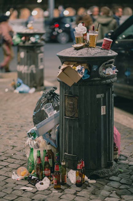 A black, cylindrical public waste bin with a domed lid is situated on a cobblestone pavement in an urban area. The bin is overflowing with various types of rubbish, including crumpled paper cups, beverage bottles, and food containers, some of which are placed on the lid or leaning against the sides of the bin. Surrounding the base of the bin, there are additional bottles, crumpled paper, and discarded items strewn across the ground, with a black plastic trash bag nearby. In the background, blurred vehicles and pedestrians are visible, indicating a busy street scene. The environment suggests an outdoor setting with natural daylight, and the scene exemplifies typical litter accumulation seen in city environments, which companies like Rubbish Clearance Harrow often address through rubbish removal services to maintain cleanliness and adhere to local waste management regulations.