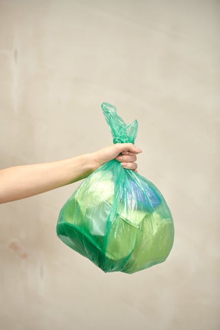 A person's hand and forearm extended into the frame from the left, holding a brightly coloured plastic rubbish bag with a knotted top. The bag is semi-transparent, showing the contents inside, and has a greenish hue with some blue areas. The background is a plain, neutral-toned wall with no additional objects or environmental details visible, typical of an indoor setting for waste disposal or rubbish collection. The scene suggests an act of disposing waste, possibly associated with private rubbish removal services like those provided by Rubbish Clearance Harrow, emphasizing the handling of domestic or commercial waste in a straightforward, professional manner.