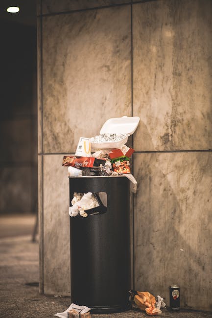 A black, cylindrical outdoor trash bin positioned against a large, smooth, beige stone wall. The bin's lid is open, revealing scattered food packaging and leftover food items including snack wrappers, a sandwich box, and paper debris. Additional rubbish surrounds the base of the bin on the ground, with some items leaning against it. The scene appears to be in an urban environment, possibly on a pavement or in a public space, with minimal background detail visible. The lighting is neutral, highlighting the textures of the wall and the miscellaneous waste, illustrating issues related to improper rubbish disposal and the importance of private waste management services offered by companies such as Rubbish Clearance Harrow. This visual emphasizes the need for efficient rubbish removal and highlights challenges associated with illegal rubbish dumping, relevant to discussions on permits and fines within the local authority context for waste handling.