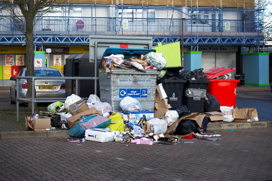 A black, cylindrical outdoor trash bin positioned against a large, smooth, beige stone wall. The bin's lid is open, revealing scattered food packaging and leftover food items including snack wrappers, a sandwich box, and paper debris. Additional rubbish surrounds the base of the bin on the ground, with some items leaning against it. The scene appears to be in an urban environment, possibly on a pavement or in a public space, with minimal background detail visible. The lighting is neutral, highlighting the textures of the wall and the miscellaneous waste, illustrating issues related to improper rubbish disposal and the importance of private waste management services offered by companies such as Rubbish Clearance Harrow. This visual emphasizes the need for efficient rubbish removal and highlights challenges associated with illegal rubbish dumping, relevant to discussions on permits and fines within the local authority context for waste handling.