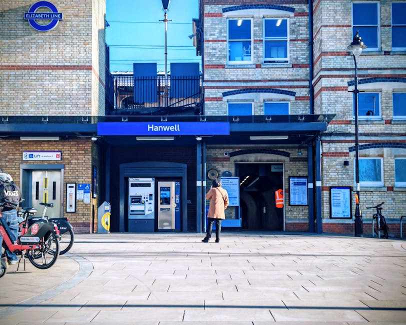 A woman in a beige coat and dark trousers stands on the paved sidewalk in front of Hanwell station, part of the Elizabeth Line, with two bicycles parked on the left side. The station entrance features a blue sign with white lettering, multiple ticket machines, and an automated barrier. The building surrounding the station is constructed from brick, with a mix of light and dark tones, and has several windows, some with white frames, and a small balcony with black railings above. A street lamp is mounted on the right side of the building next to a small bicycle rack. Bright sunlight illuminates the scene, casting shadows across the pavement, and the environment appears clean and well-maintained, representative of an urban public transport hub. The presence of bicycles and ticket machines indicates a multimodal transit point, while the station's architecture and signage imply a focus on accessible, independent travel options within the area. Rubbish clearance services by Rubbish Clearance Harrow could be relevant for maintaining such busy public spaces, especially in terms of waste management in urban transit environments.