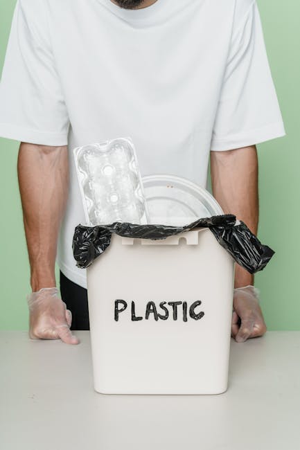 A person wearing a white T-shirt and transparent disposable gloves is holding a white plastic bin with the word 'PLASTIC' written on it in black. The bin contains a black plastic rubbish bag and an empty plastic blister pack, which is leaning against the inner edge of the bin. The setup is placed on a flat surface with a light green background. The image visually relates to waste management and rubbish collection, emphasizing the segregation and disposal of plastic waste, a service that Rubbish Clearance Harrow offers. The scene highlights the importance of organized waste separation for environmentally responsible rubbish removal, aligning with the context of independent collection or private disposal services related to rubbish removal in Harrow.