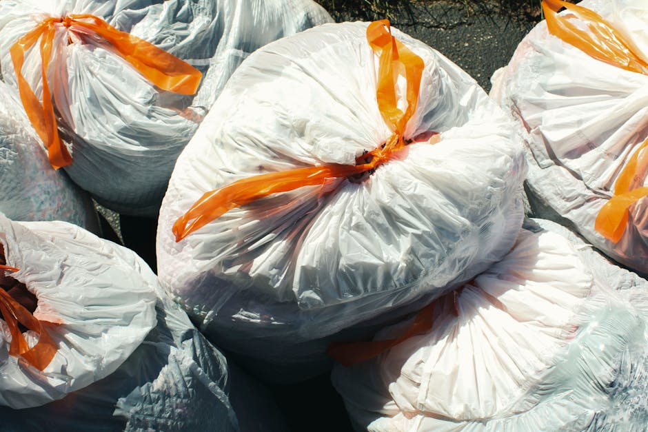 A pile of overflowing rubbish bags, cardboard boxes, and miscellaneous waste items is accumulated on a paved sidewalk in an urban setting, near a parked silver car with a yellow registration plate. The waste includes black plastic rubbish bags, flattened cardboard boxes, plastic packaging, and paper materials, some crumpled or torn, with a mix of colors such as black, brown, white, and green. The pile is situated beside metal railings that separate the sidewalk from a small parking area. In the background, there are commercial premises with storefronts, including signs and window displays, and a building under renovation or construction, covered in scaffolding and blue protective netting. The environment appears to be during daylight hours under natural lighting, with a tree partially visible on the left side. This scene exemplifies the type of waste collection that Rubbish Clearance Harrow provides as an alternative to official rubbish disposal, illustrating a typical scenario where private waste removal services are required for on-site clearance outside standard council collections.