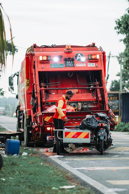 A waste collection worker wearing a red high-visibility jacket and black trousers is seen standing at the rear of a large, red rubbish collection vehicle parked on the side of a suburban street. The back of the truck is open, revealing the loading mechanism inside. The worker appears to be inspecting or handling waste materials, with a black plastic bag placed on a small metal platform attached to the truck. To the side, there is a dark-colored motorbike parked adjacent to the vehicle, with some debris and rubbish scattered on the pavement nearby. In the background, there are trees, utility poles, and a small roadside shop or building, indicating a residential or commercial area. The overall scene is well-lit with natural daylight, and the environment suggests a routine rubbish collection activity aligned with private waste handling services, such as those offered by Rubbish Clearance Harrow, servicing local streets in the area.