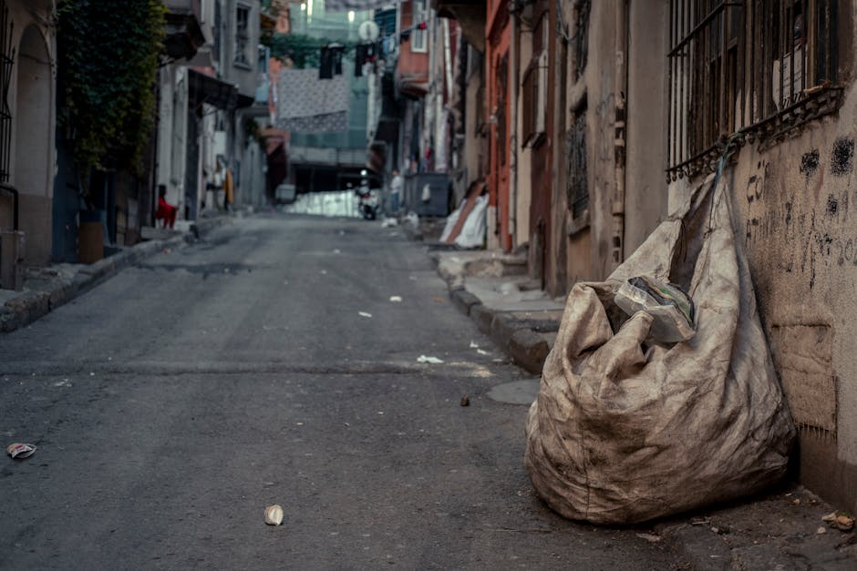 A large, worn, beige fabric rubbish bag is placed on the uneven pavement of a narrow urban street, leaning against a weathered concrete wall with visible graffiti and metal security bars. The bag appears partially filled, with some plastic bottles protruding from the top, indicating it contains household waste. Surrounding the bag are scattered pieces of trash and small debris, with the street extending into the distance, lined with closely packed buildings featuring various textures, colors, and open windows. The environment appears somewhat neglected, with signs of wear on the buildings and street surface showing cracks and irregularities. In the background, garments and laundry are hung on lines stretching between buildings, and a few miscellaneous objects, such as a red cloth and a bicycle, are visible further along the street. The scene is lit by natural daylight, creating a neutral, utilitarian atmosphere typical of private waste disposal in urban alleyways, and subtly relates to independent rubbish removal services offered by Rubbish Clearance Harrow.