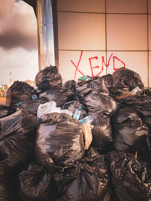 A waste collection worker wearing a red high-visibility jacket and black trousers is seen standing at the rear of a large, red rubbish collection vehicle parked on the side of a suburban street. The back of the truck is open, revealing the loading mechanism inside. The worker appears to be inspecting or handling waste materials, with a black plastic bag placed on a small metal platform attached to the truck. To the side, there is a dark-colored motorbike parked adjacent to the vehicle, with some debris and rubbish scattered on the pavement nearby. In the background, there are trees, utility poles, and a small roadside shop or building, indicating a residential or commercial area. The overall scene is well-lit with natural daylight, and the environment suggests a routine rubbish collection activity aligned with private waste handling services, such as those offered by Rubbish Clearance Harrow, servicing local streets in the area.