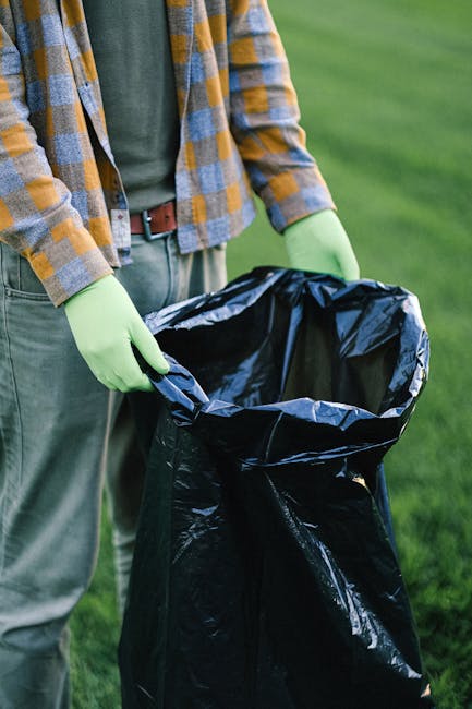 A person wearing a yellow, grey, and blue checkered shirt over a green T-shirt and grey trousers, with a black belt, is standing outdoors on a grassy area during daytime. They are wearing bright green rubber gloves and holding open a large black plastic rubbish bag, which is partially transparent and lined with a smaller silver bag inside. The background features a well-maintained green lawn, indicating a clean, outdoor environment suitable for waste collection. The scene appears to depict someone engaged in rubbish removal or clearance activities, aligning with services offered by Rubbish Clearance Harrow for private waste disposal or on-site clearance, with no visible waste inside the bag yet but prepared for rubbish collection or disposal.