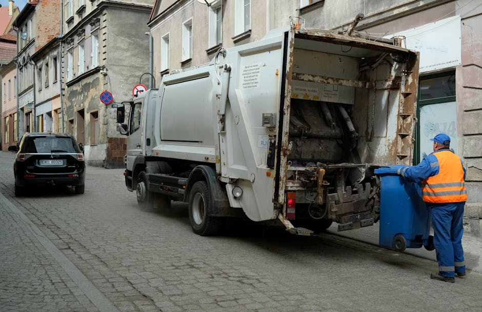 A waste collection vehicle parked on a narrow cobblestone street in an urban area, with a worker in a blue uniform and high-visibility orange vest disposing of a blue wheeled bin into the rear hopper of the garbage truck. The truck's metal rear loading section shows signs of rust and wear, with hydraulic mechanisms visible for lifting and emptying bins. On the left side, a black compact car is parked close to the curb, and behind it, there are older residential buildings with beige and grey facades, some showing signs of age and patchwork repairs. The street is lined with a mix of small shopfronts and residential windows, with street signs indicating parking restrictions. The scene is lit with natural daylight, offering a clear view of the urban environment and the process of on-site waste collection aligned with private rubbish removal services such as those provided by Rubbish Clearance Harrow.