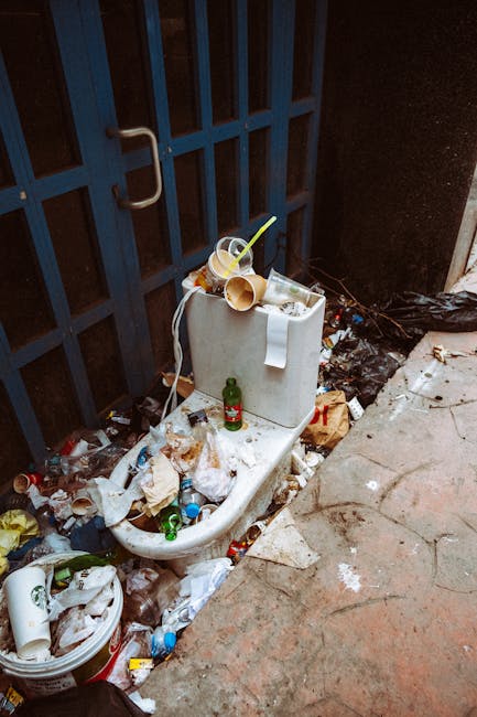 The image displays a white ceramic toilet that has been abandoned outside in an area adjacent to a black metal gate with vertical bars and a silver handle. The toilet is cluttered with various discarded items, including plastic bottles, paper waste, and food containers, some of which are partially crushed or crumpled. On top of the toilet tank, there are two toppled paper cups with yellow and white straws, along with a small green glass bottle standing upright. Surrounding the base of the toilet, there is a significant amount of rubbish scattered across the uneven concrete ground, including plastic bags, wrappers, and additional beverage containers. To the left of the toilet, a white plastic bucket overflows with more waste, indicating environmental neglect. The overall scene suggests an informal or neglected space, where private waste or rubbish has been dumped, highlighting the importance of proper rubbish removal services such as those offered by Rubbish Clearance Harrow for alternative waste handling outside standard municipal collection.